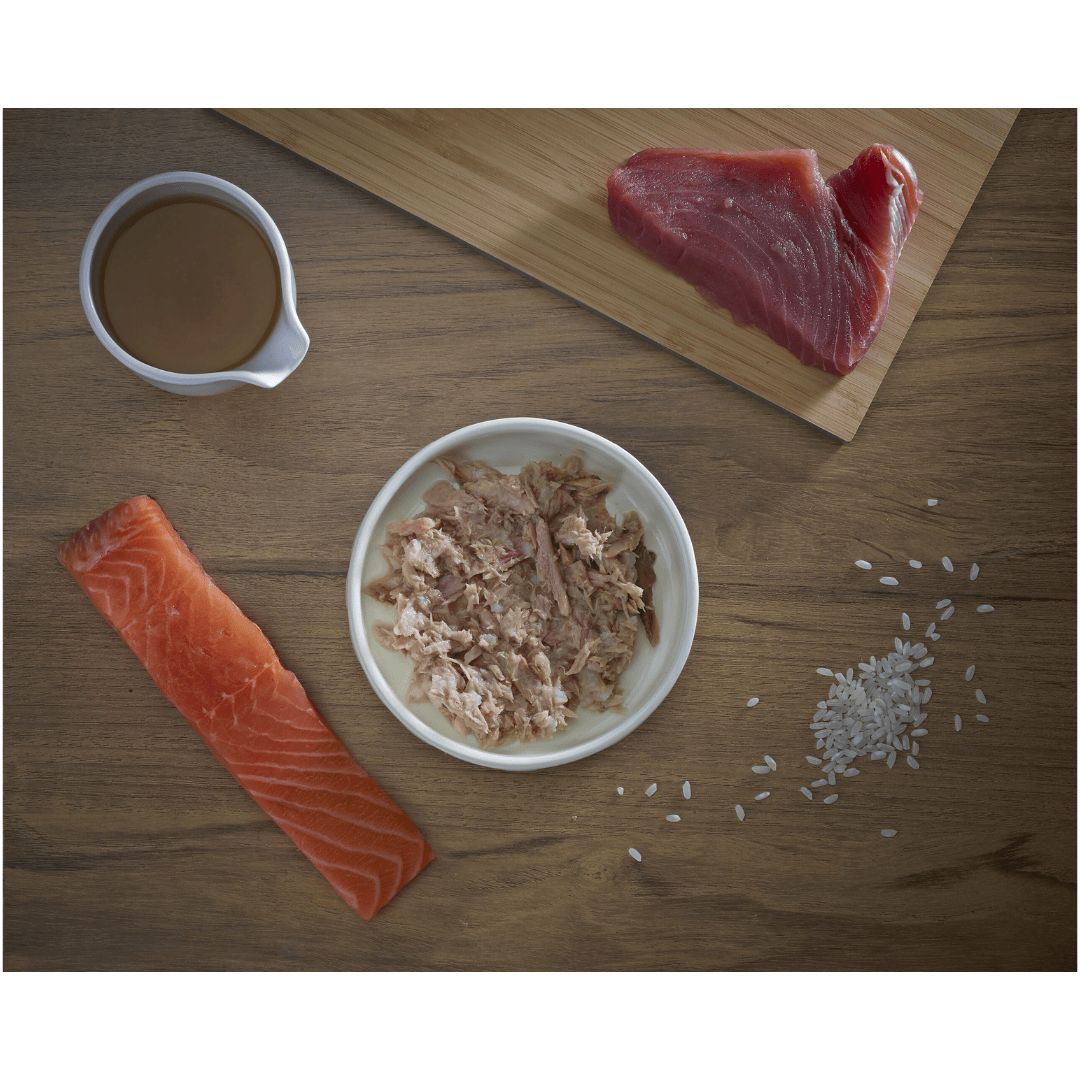 Some fresh ingredients, tuna, salmon, broth and rice. In the middle of these, there is a bowl of Fish 4 Cats Tuna & Salmon served in a bowl.