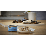 The Fish 4 Cats Sardine & cat food served on a plate with a soft, fish consistency, there are chunks of mussels too. The tin is beside the bowl. There is Sardines and mussels on display in the background.